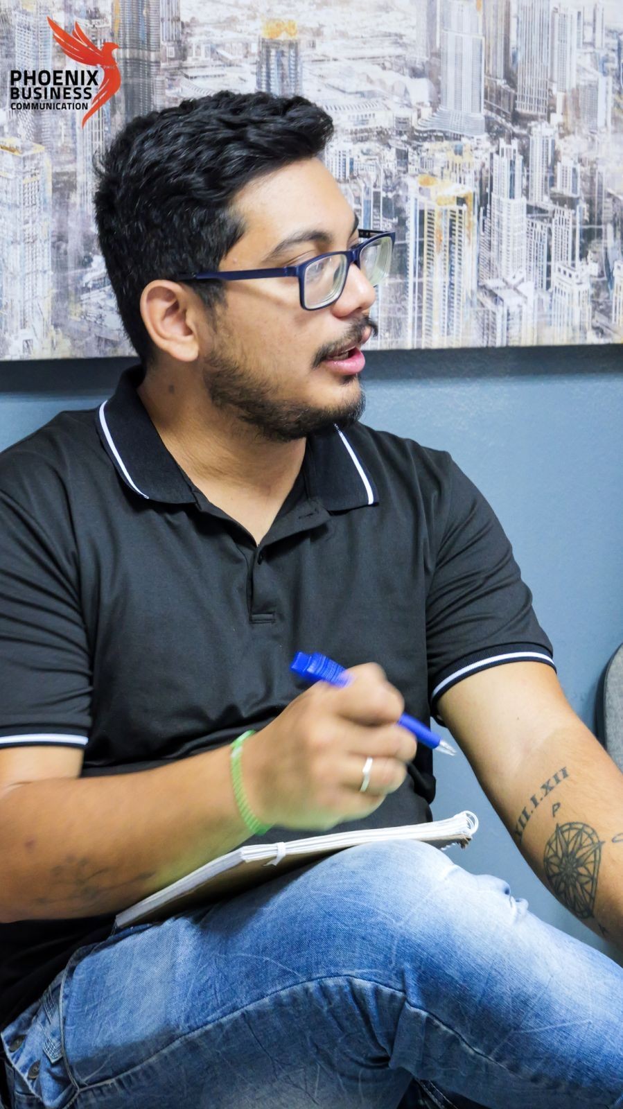 Man in glasses holding a pen and notebook, sitting in front of a cityscape painting.