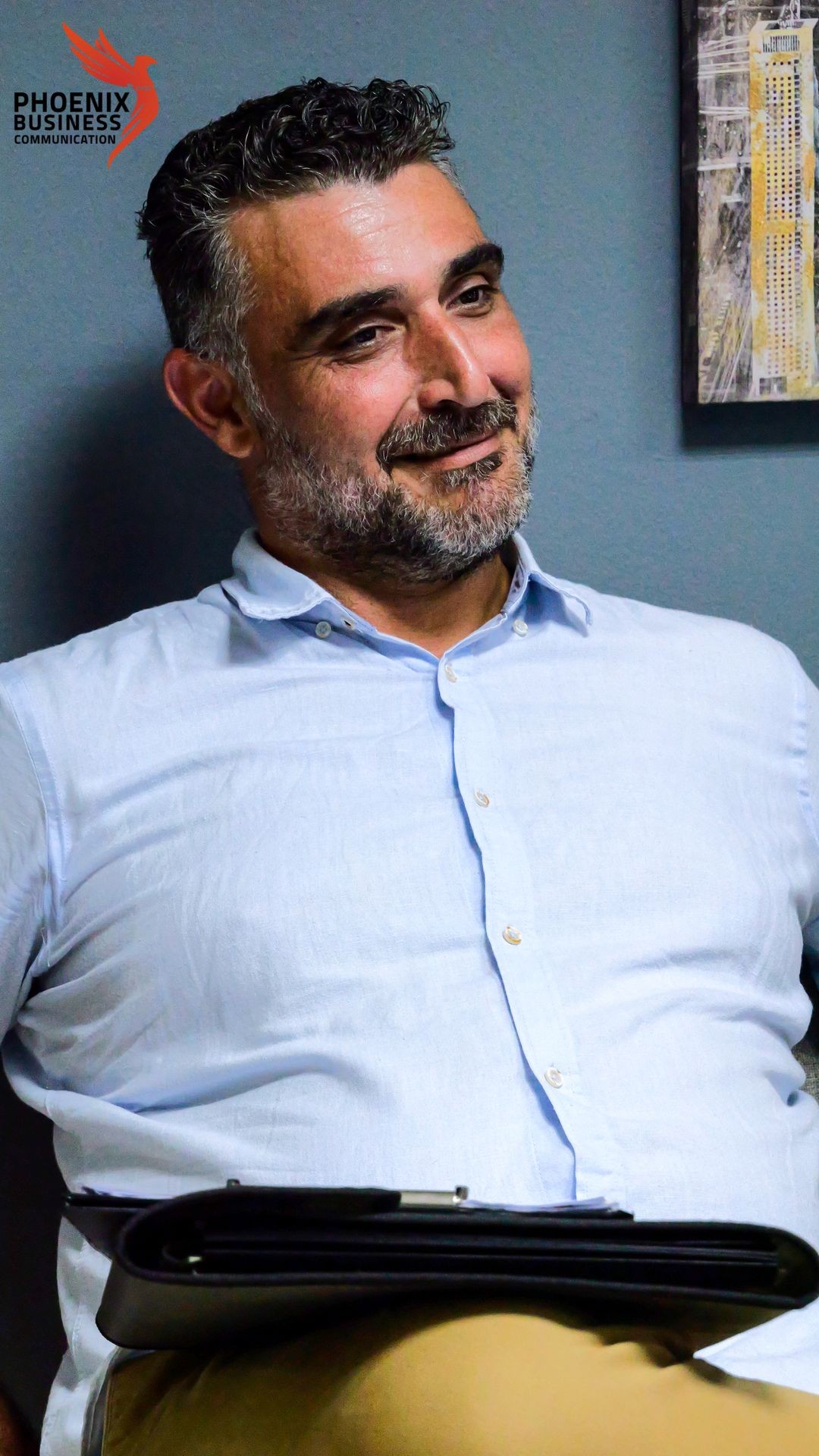 Man in a light shirt smiles while seated indoors, holding a folder.