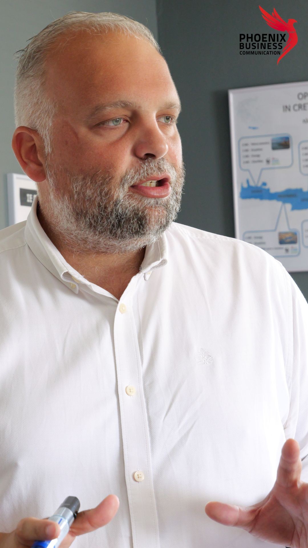 Man speaking in front of an office setting, wearing a white shirt and holding a marker.