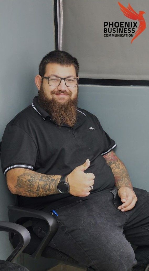 Bearded man with tattoos sitting and giving a thumbs up, wearing glasses and a black shirt.