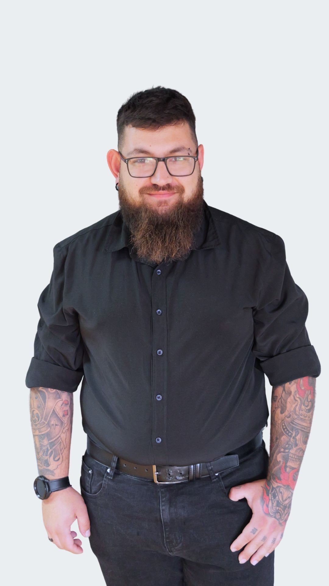 Man in black shirt and glasses with tattoos on arms, standing against pale background.