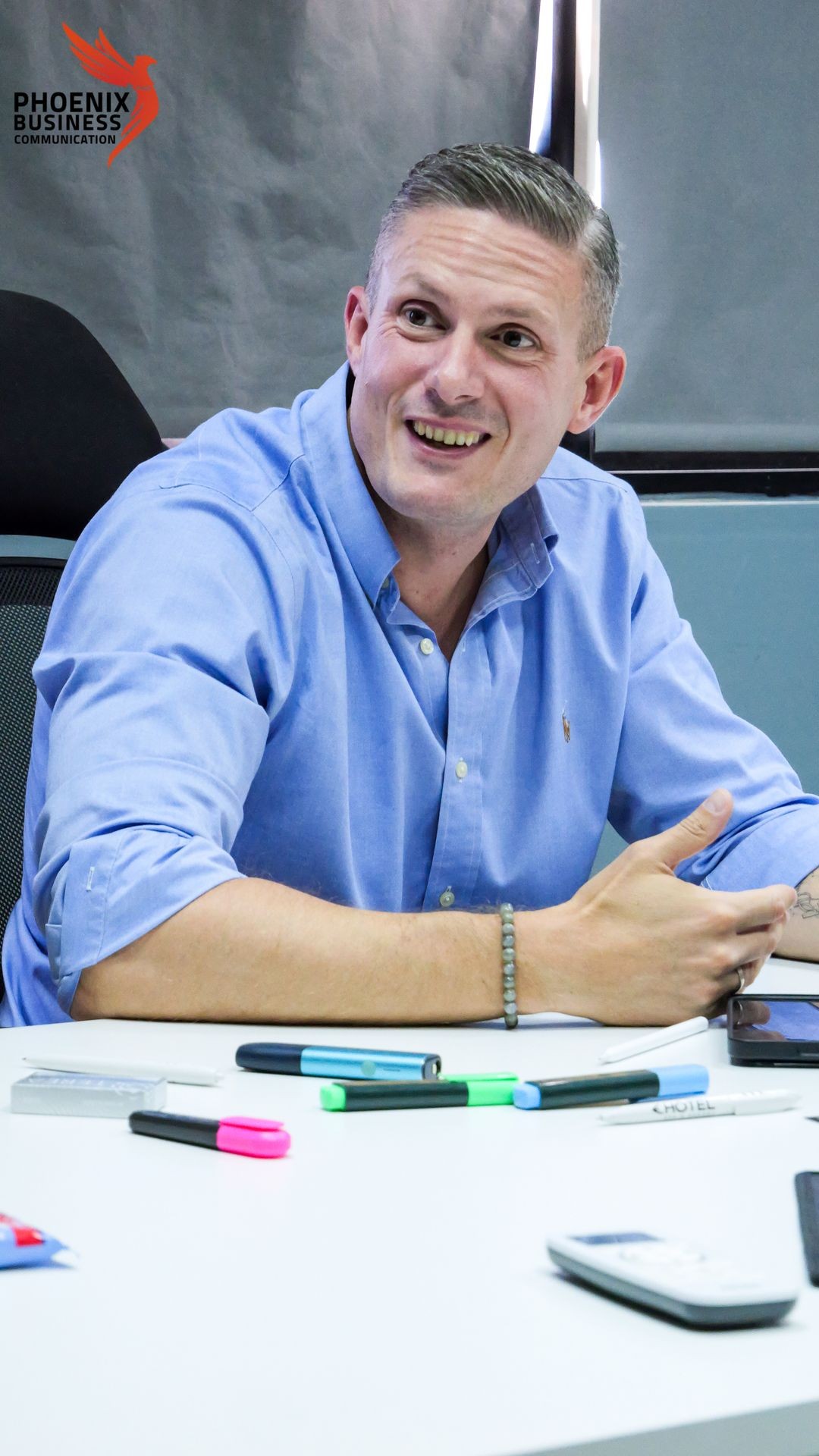 Man in a blue shirt smiling at a desk with colorful markers and pens.