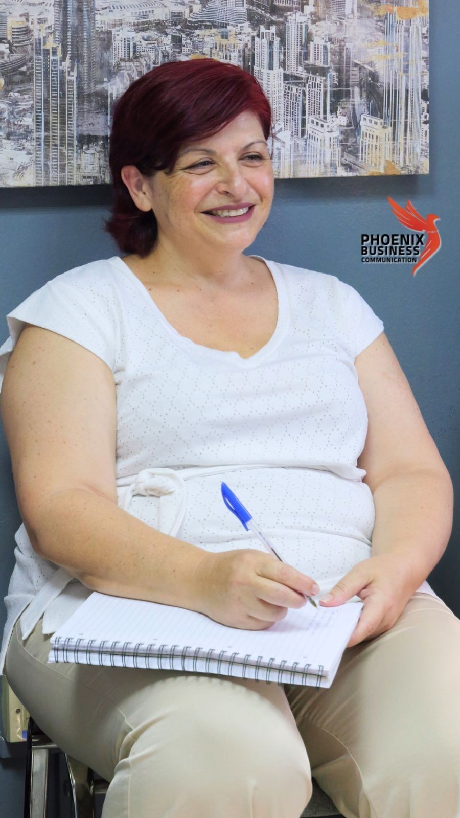 Woman with red hair smiling and writing in a notebook, seated indoors near a cityscape painting.