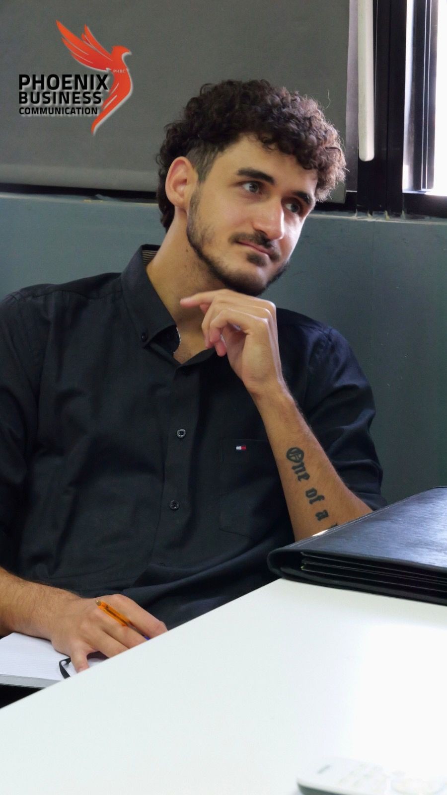 Man in black shirt sitting thoughtfully with a pen, logo of Phoenix Business Communication in the background.