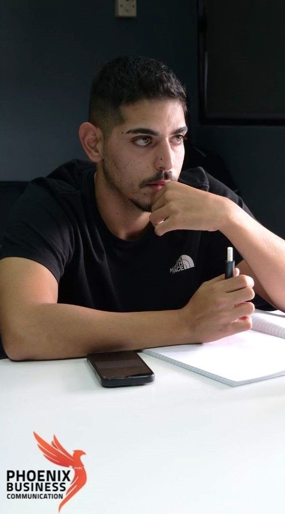 Man in black shirt holding pen, thinking, with phone and notebook on table.