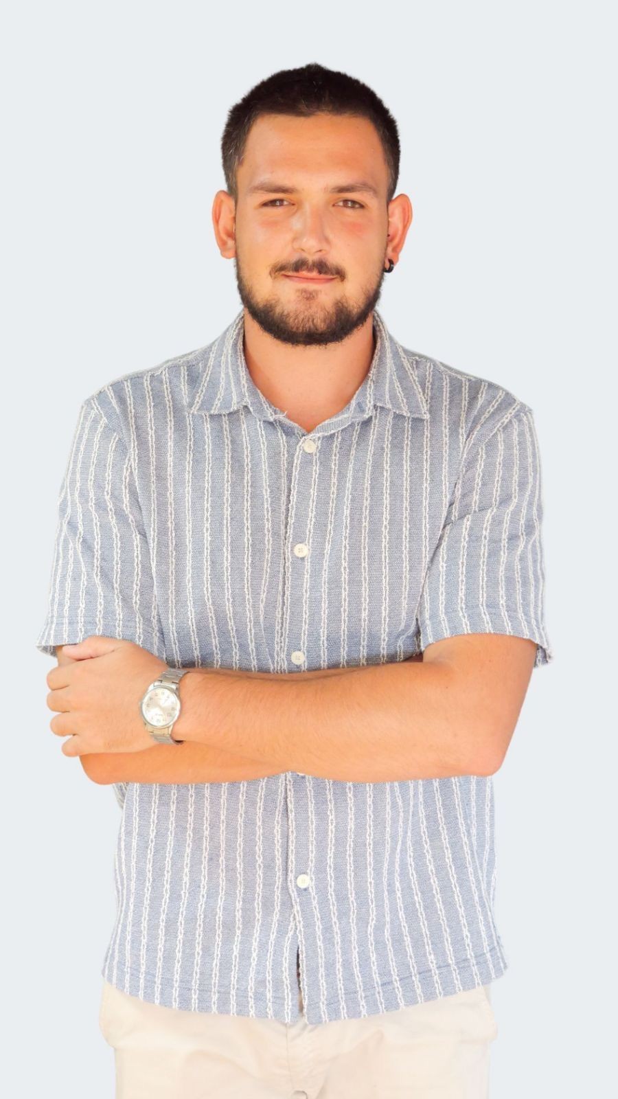Man in a striped shirt with crossed arms and a watch, standing against a plain background.